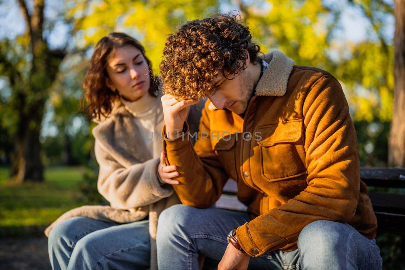 Young couple is sitting in park on sunny day. Man is sad and woman in consoling him.