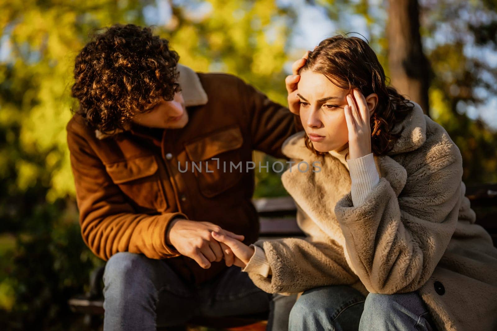 Young couple is sitting in park on sunny day. Woman is sad and man in consoling her.