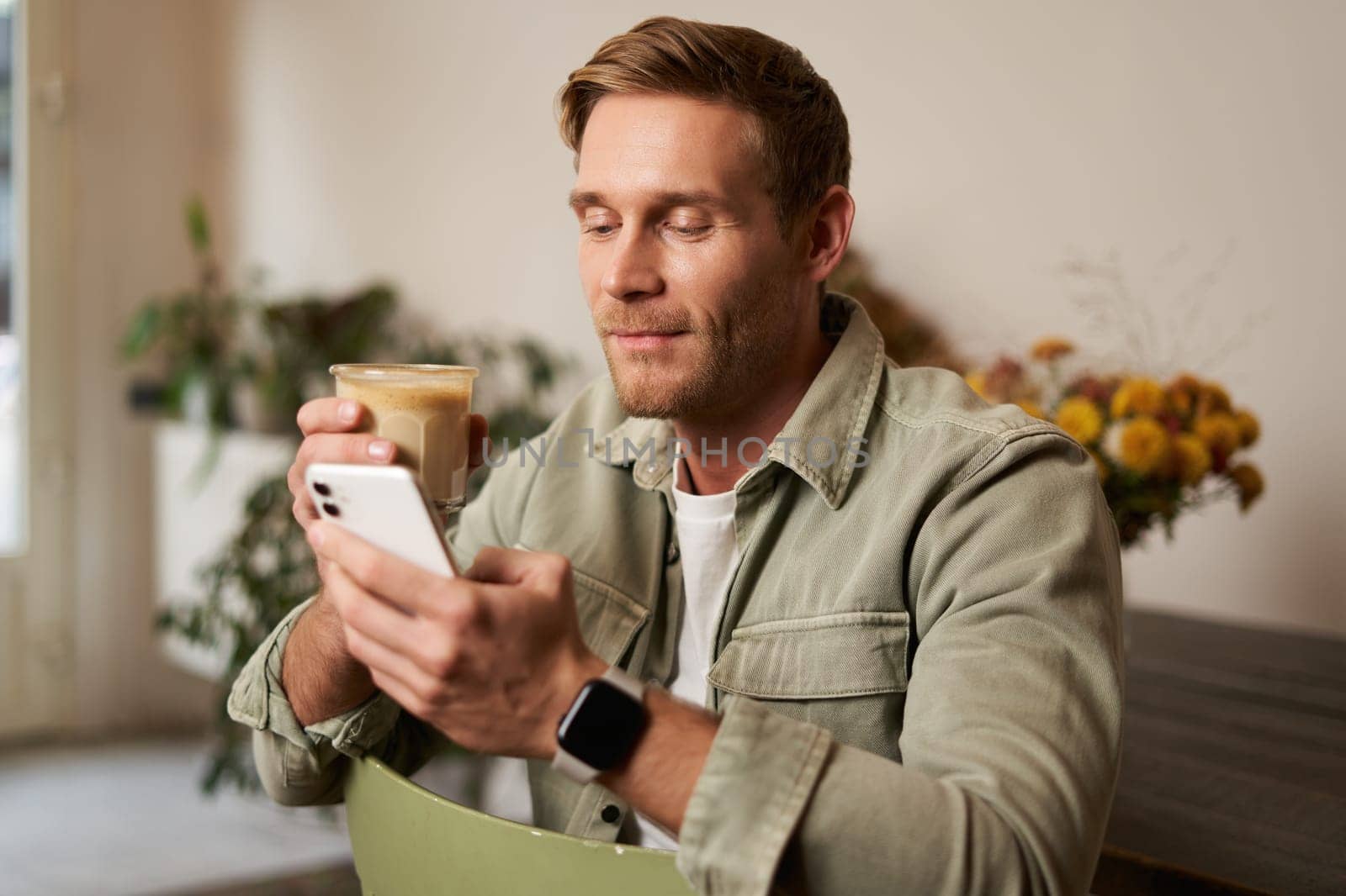 Lifestyle portrait of handsome young man, sitting in cafe, checking his phone and drinking coffee, smiling. by Benzoix