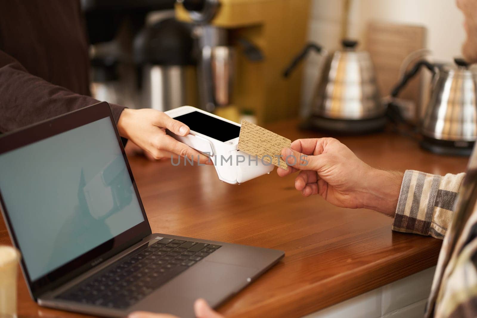 Cropped shot of male hand paying contactless for his order in coffee shop, sitting at counter with laptop, pressing card to POS terminal, working from home.