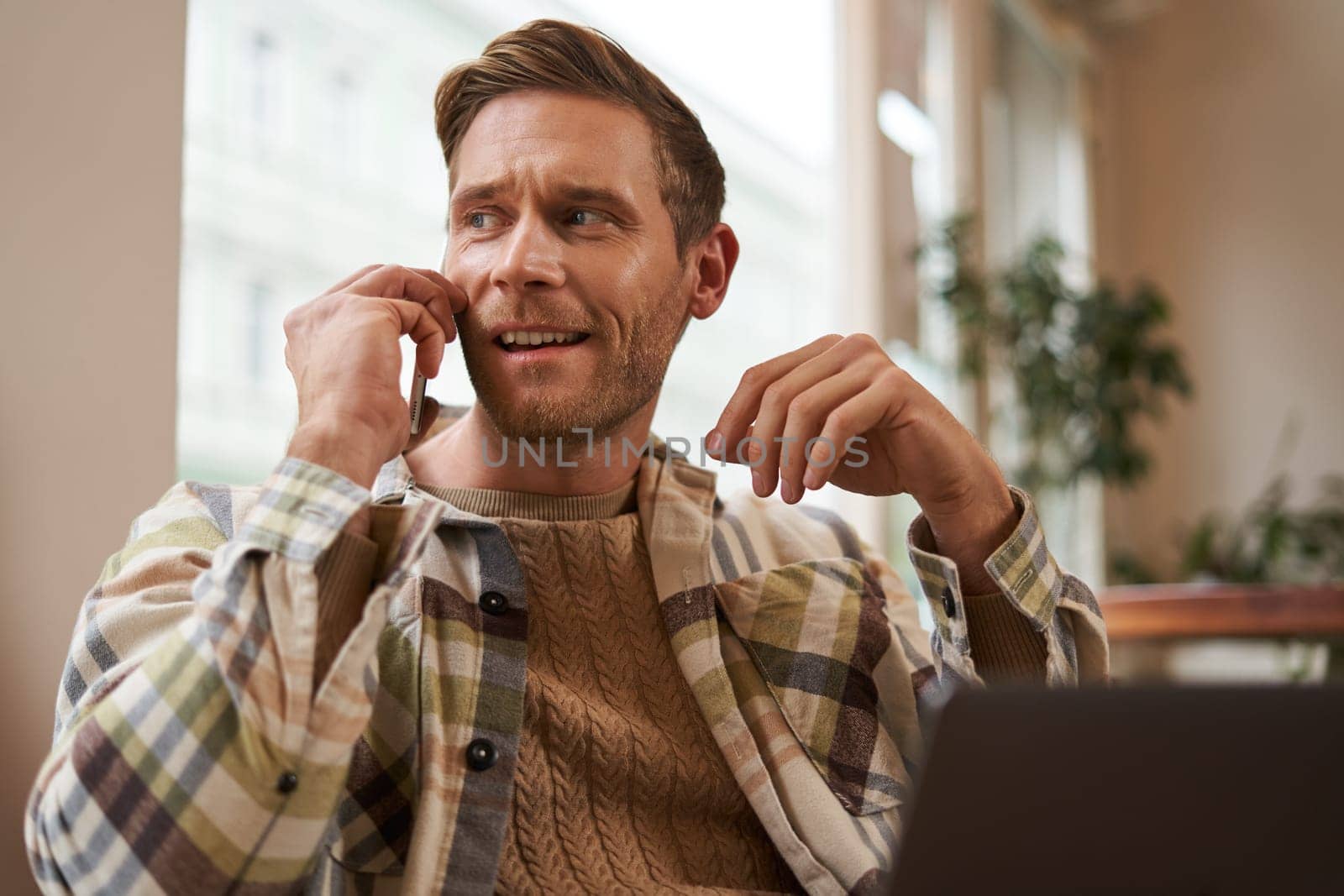 Portrait of young businessman, freelancer working remotely from coffee shop in city, sitting in chair with laptop, answer a phone call, talking on mobile to someone.