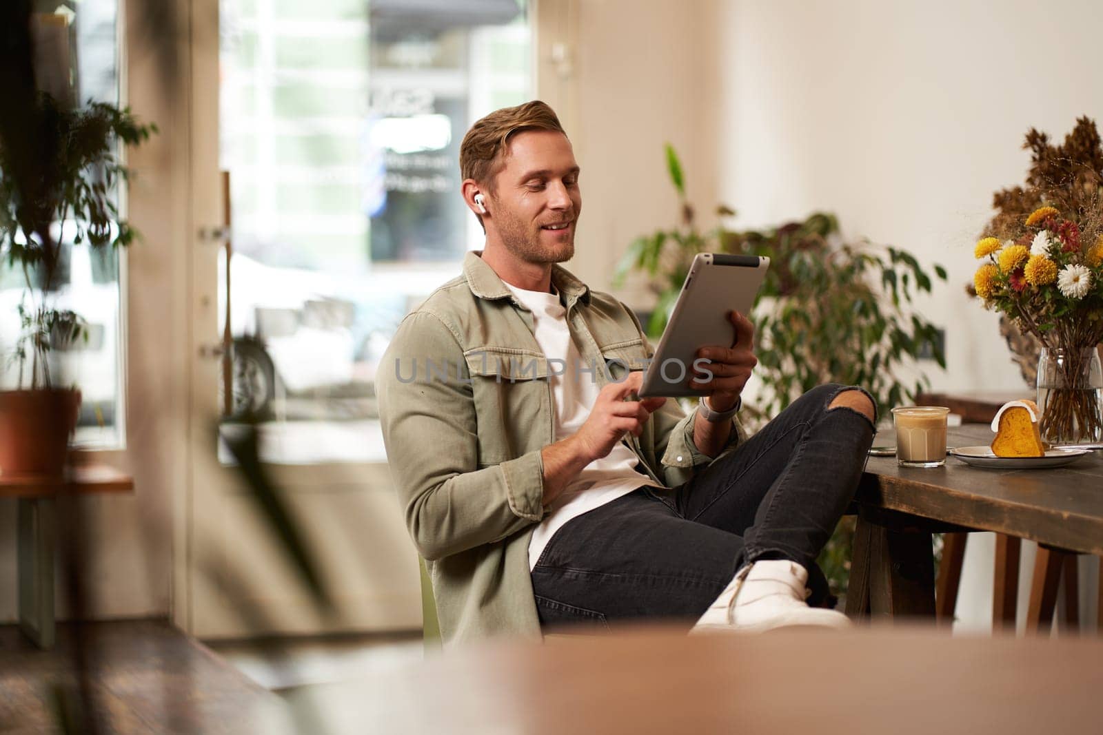 Portrait of young man sitting in cafe, spending time alone in coffee shop, reading on digital tablet, watching video, listening music in wireless headphones. Lifestyle and people concept.