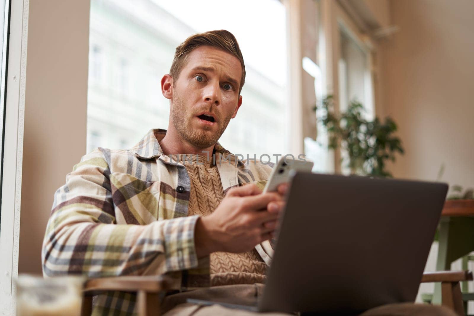 Image of man with shocked, concerned face, after reading terrible news on phone. Adult man in coffee shop, sitting in cafe with laptop, reacting worried after reading smth on smartphone.