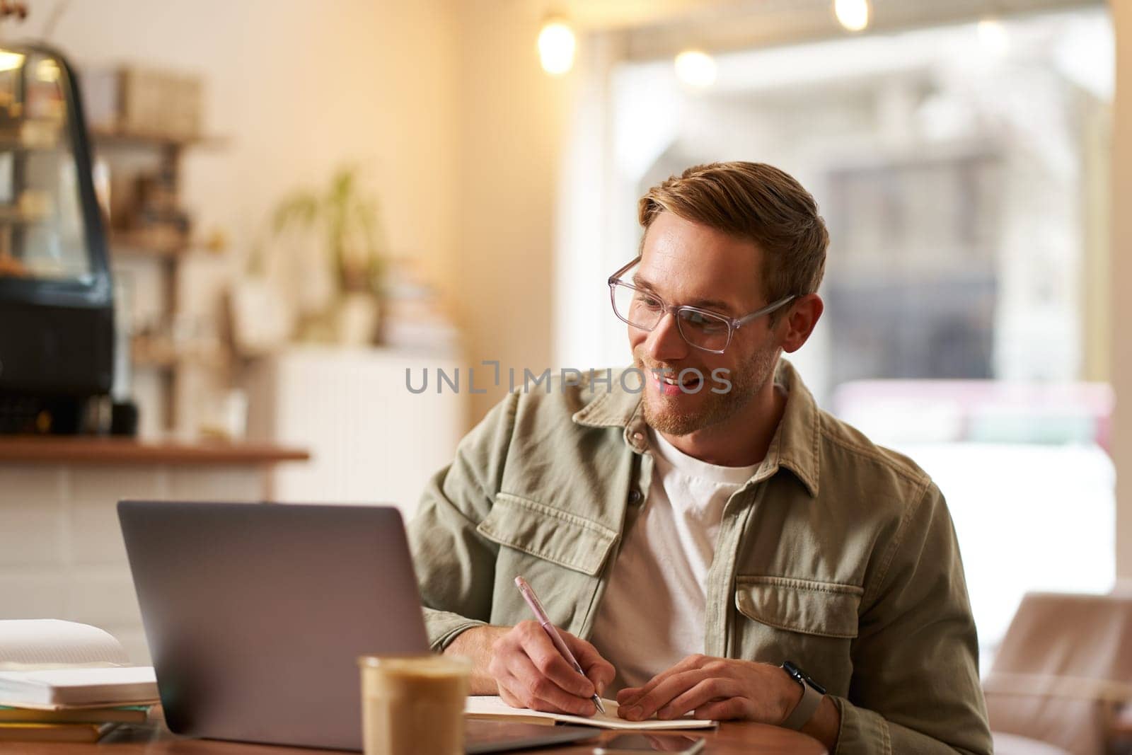 Portrait of modern man in glasses, sitting in quite empty cafe with laptop, writing down notes in notebook, studying online, remote e-learning, passing web course by Benzoix