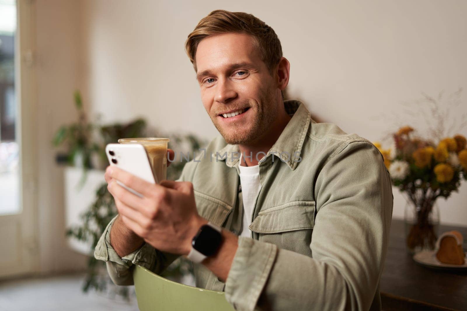 Lifestyle portrait of handsome young man, sitting in cafe, checking his phone and drinking coffee, smiling. Concept of leisure, going out