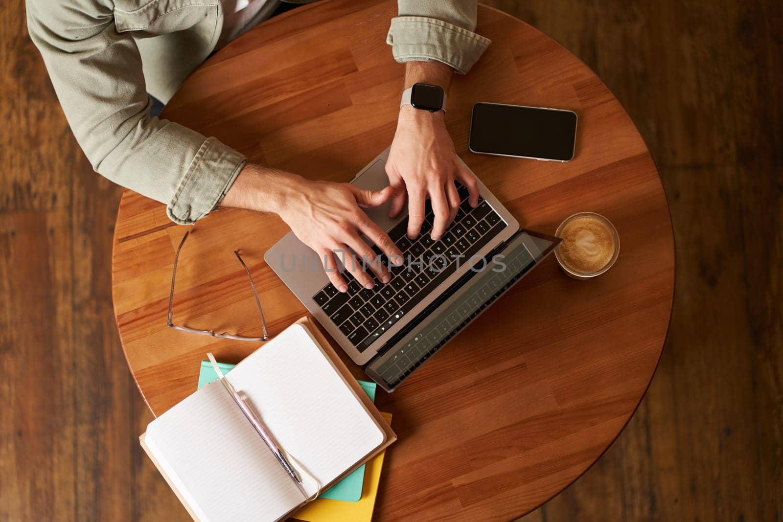 Cropped top view of male hands, man typing on laptop keyboard, working in cafe, sitting at round table in coffee shop, has phone, notebook and cup.