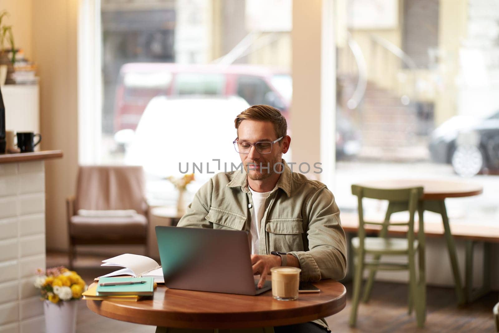 Image of young digital nomad, man in glasses sits in cafe, works from coffee shop, uses laptop in co-working space, wears glasses, drinks his beverage.