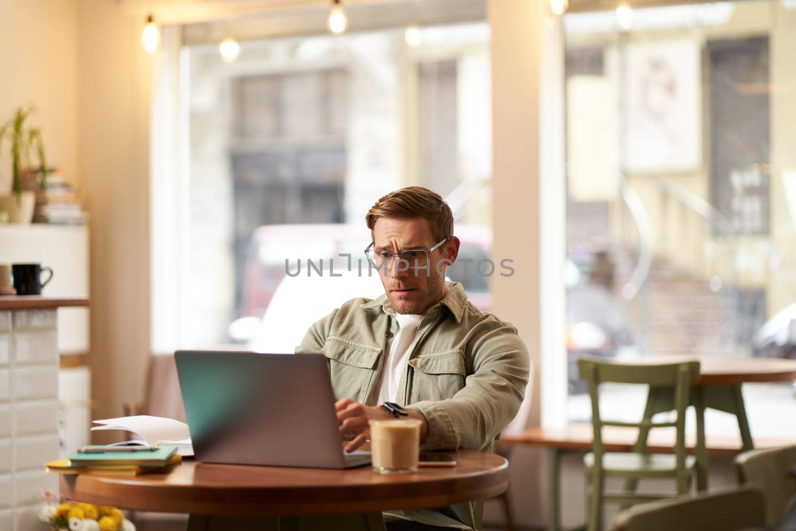 Image of young digital nomad, man in glasses sits in cafe, works from coffee shop, uses laptop in co-working space, wears glasses, drinks his beverage.
