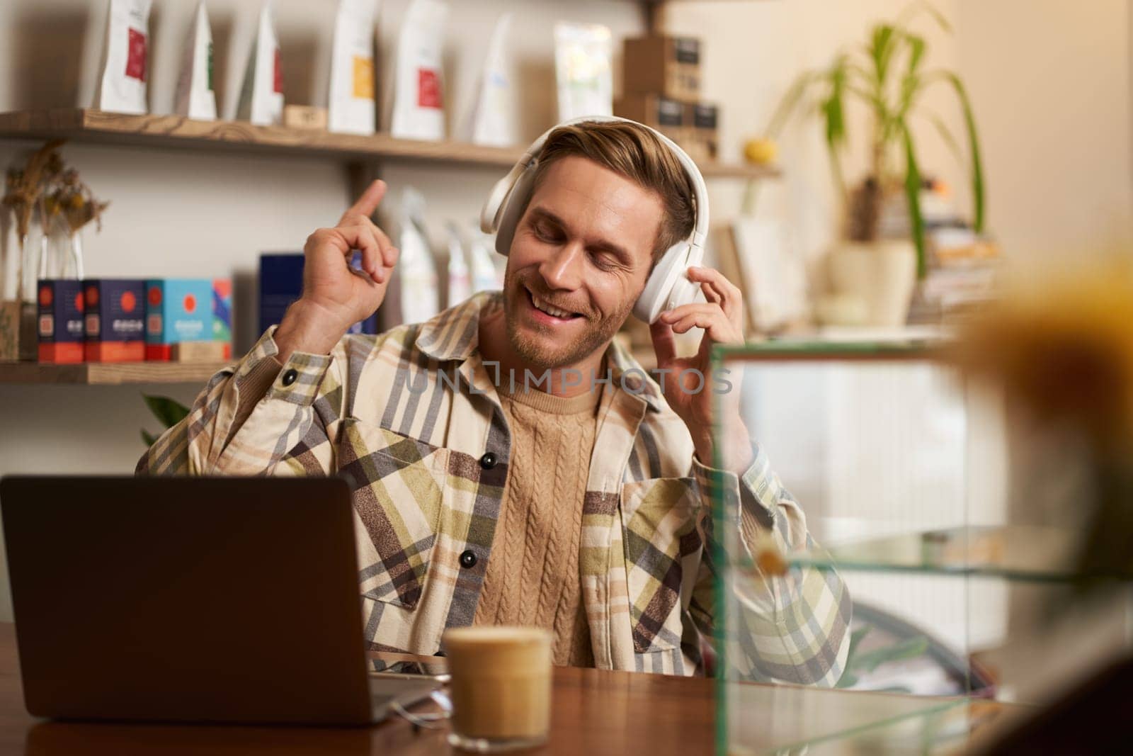 Lifestyle portrait of smiling digital nomad, man in cafe, working on laptop, listening music in wireless headphones and smiling, dancing on chair while enjoying favourite song.