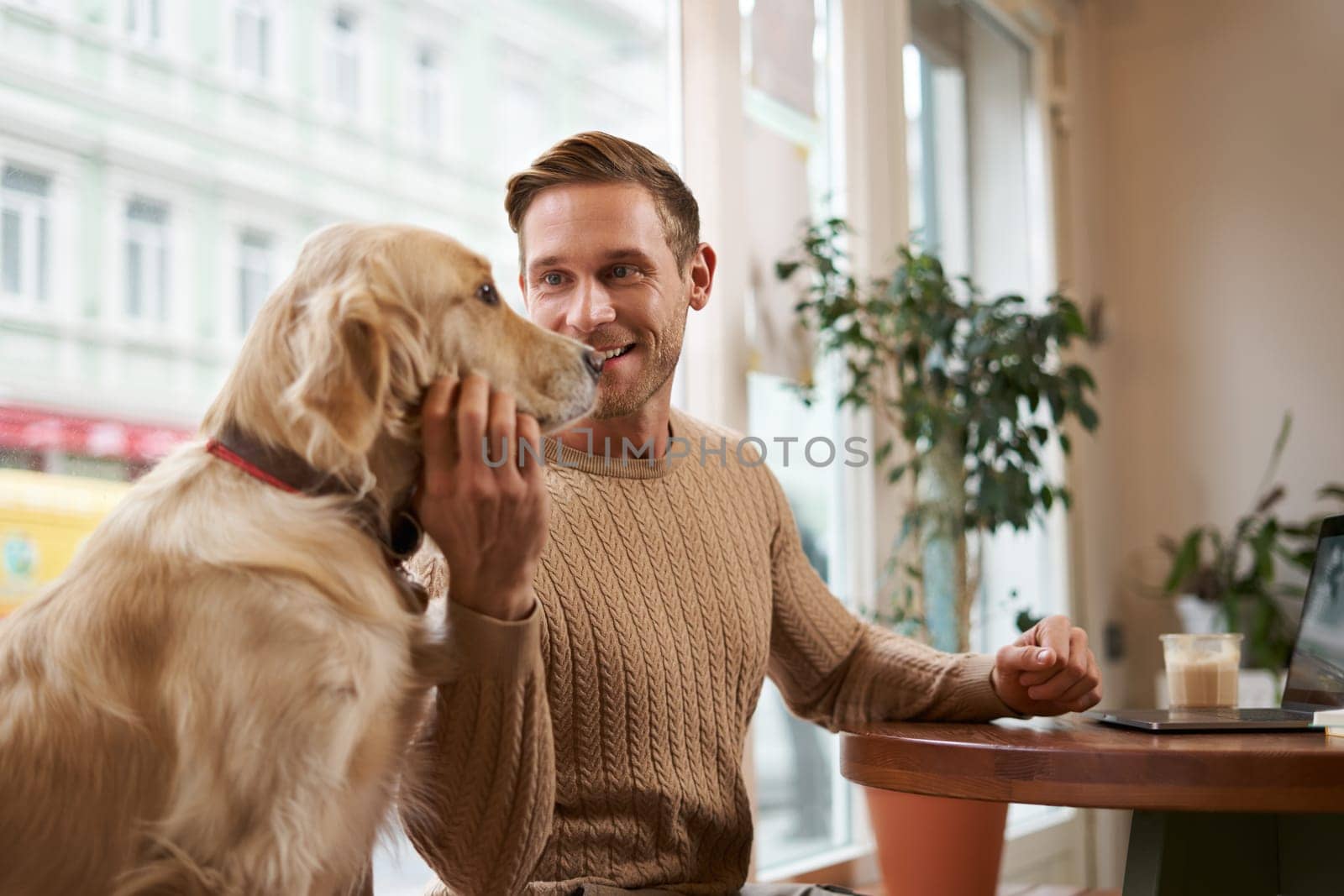 Lifestyle photo of young handsome man, entrepreneur working in a cafe on laptop while his dog sits next to him. A guy pets his golden retriever while using computer in a coffee shop.