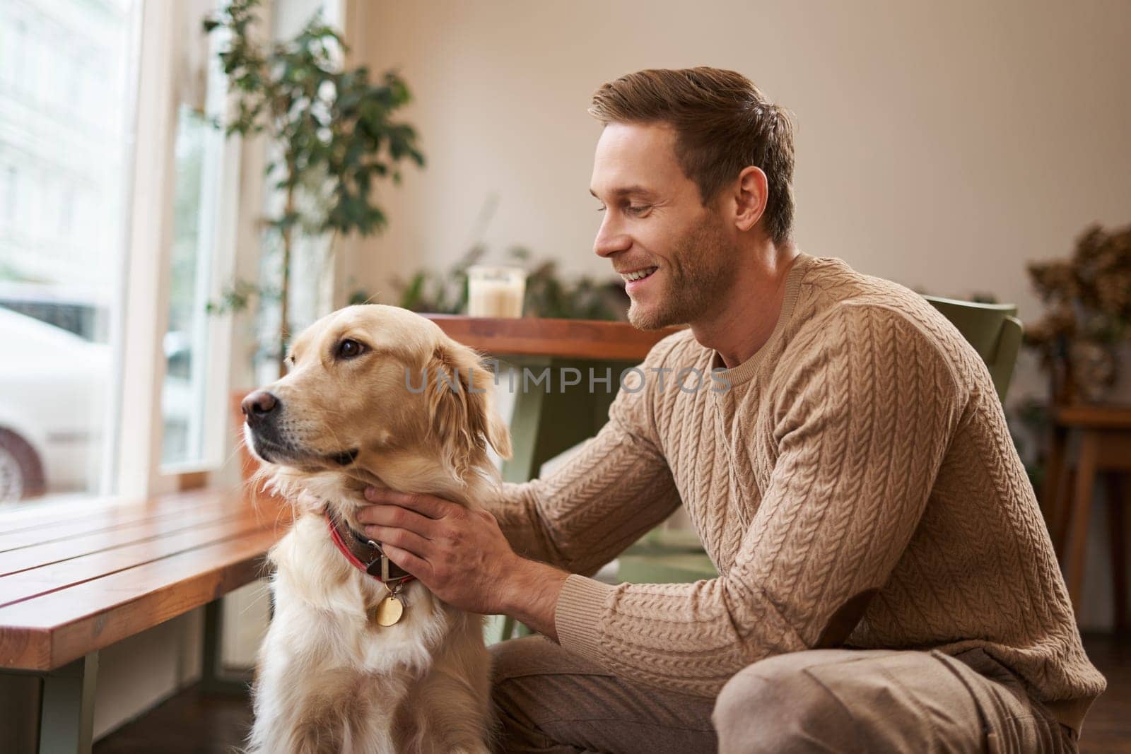 Portrait of young handsome dog-owner, spending time with his pet in a cafe, sitting indoors, looking outside window.