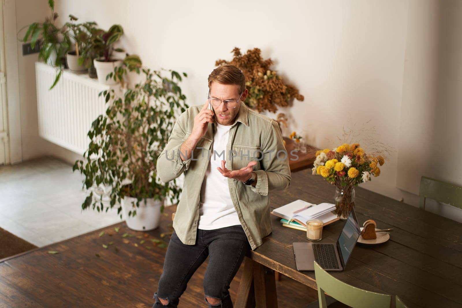 Portrait of stressed man, business manager, talking on mobile phone with concerned, nervous face expression, having a serious conversation, arguing over the telephone in office, sitting on table.