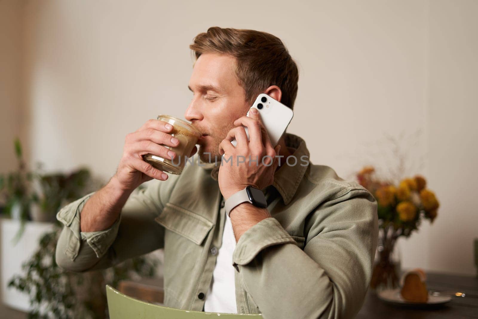 Close up portrait of handsome man enjoys his coffee, drinks cappuccino, talks on mobile phone, answers a phone call, sits in cafe.