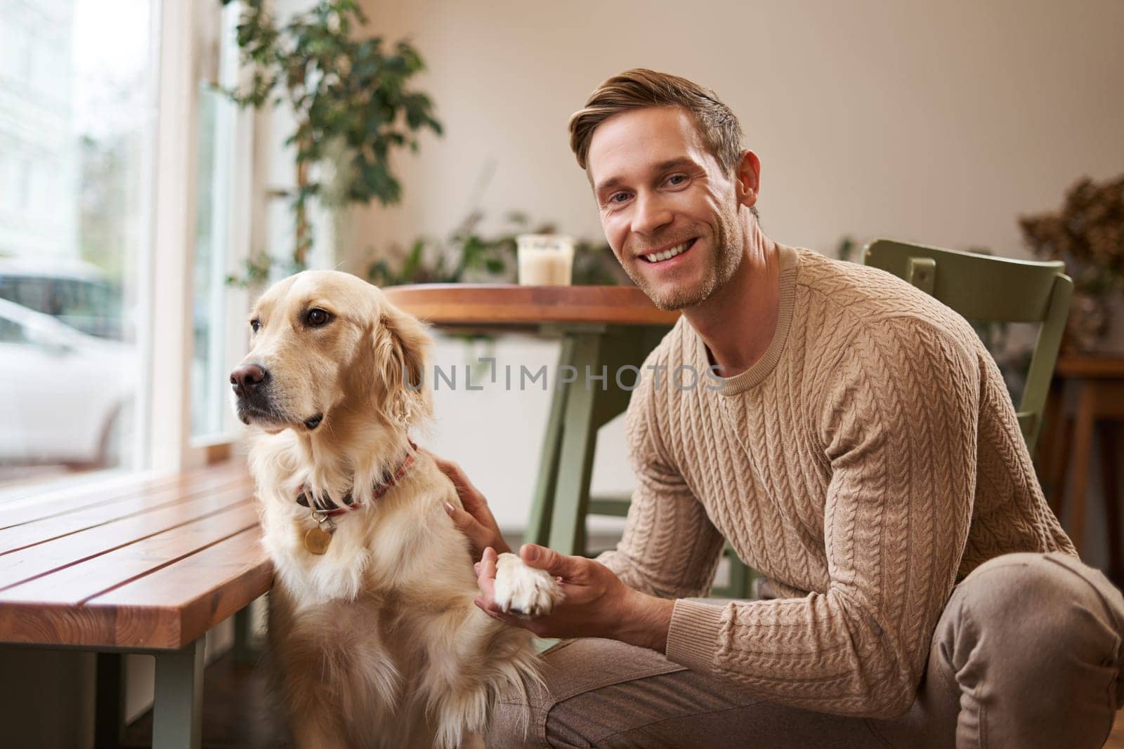 Portrait of young handsome dog-owner, spending time with his pet in a cafe, sitting indoors, looking outside window by Benzoix