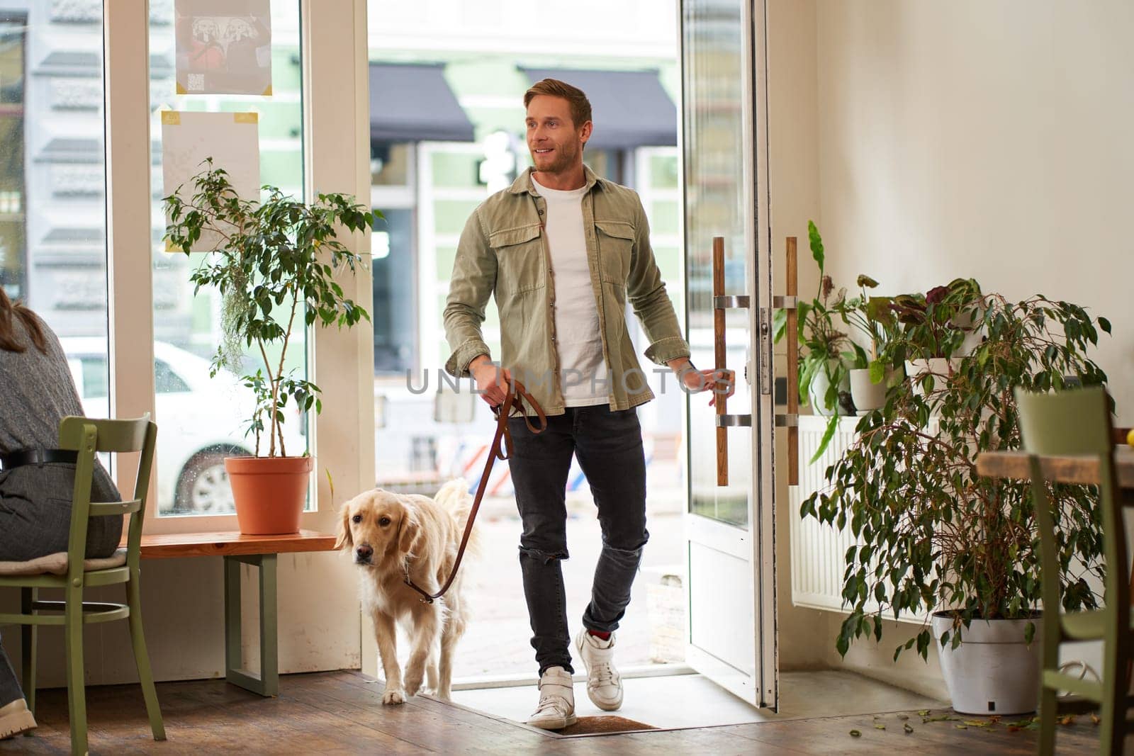 Portrait of handsome smiling young man, walking into the coffee shop with his cute dog, golden retriever on leash. Guy enters pet-friendly cafe.