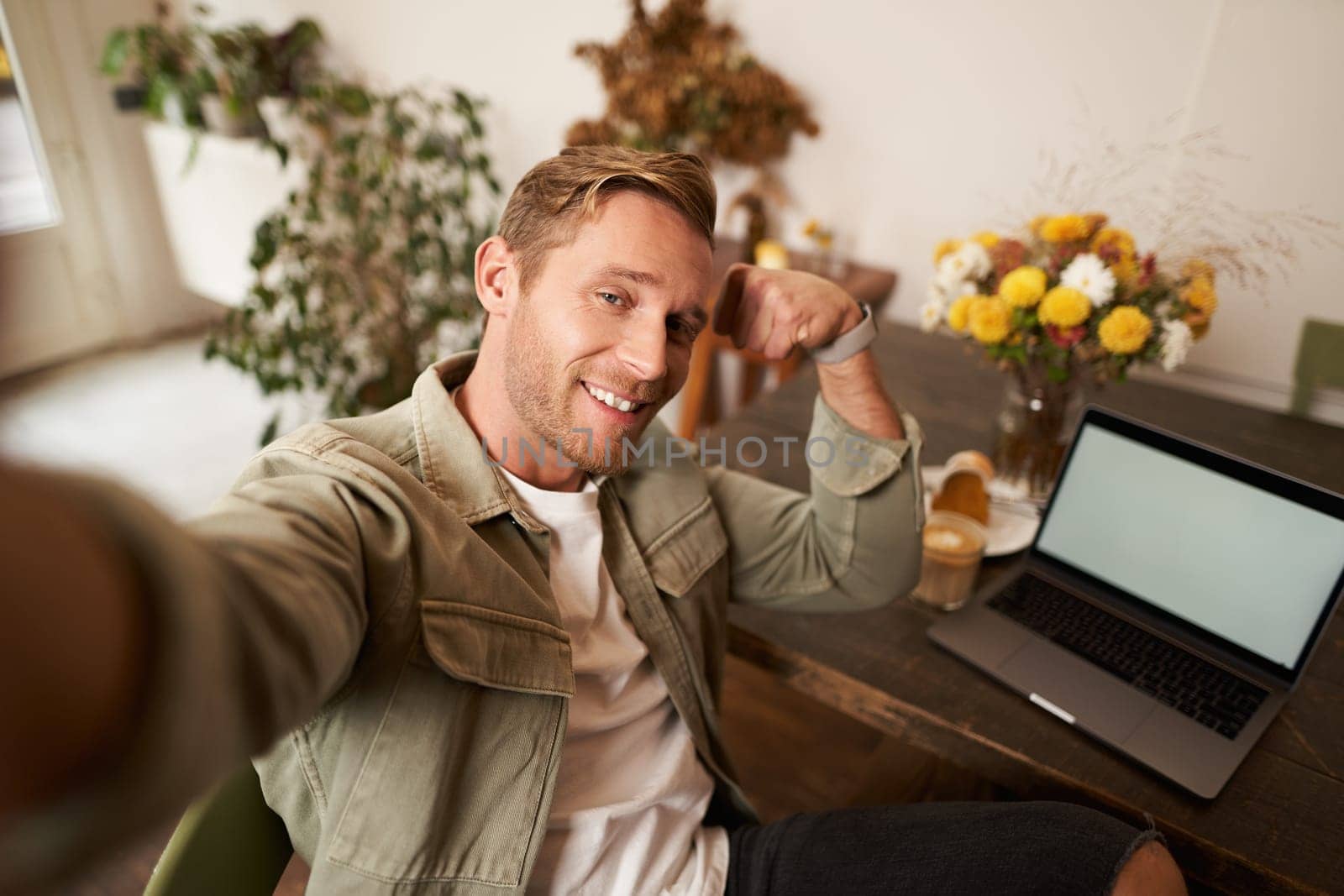 Portrait of handsome and confident man taking a selfie, flexing biceps, shows-off his muscles, sits in cafe in front of laptop, smiling at camera.
