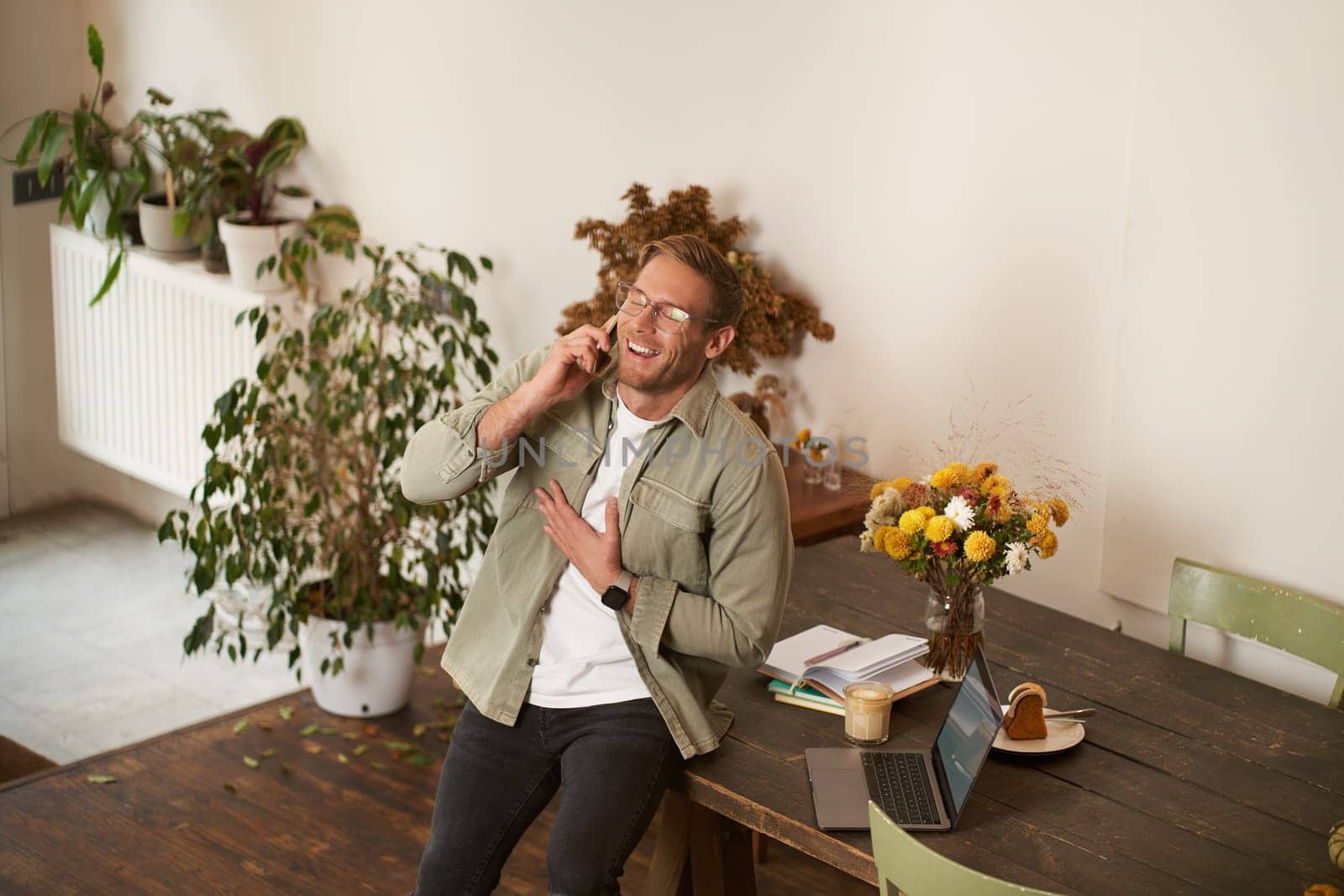 Portrait of handsome office manager, guy in co-working space, talking on mobile phone, calling someone, laughing and smiling, having pleasant conversation, sitting in office on table.