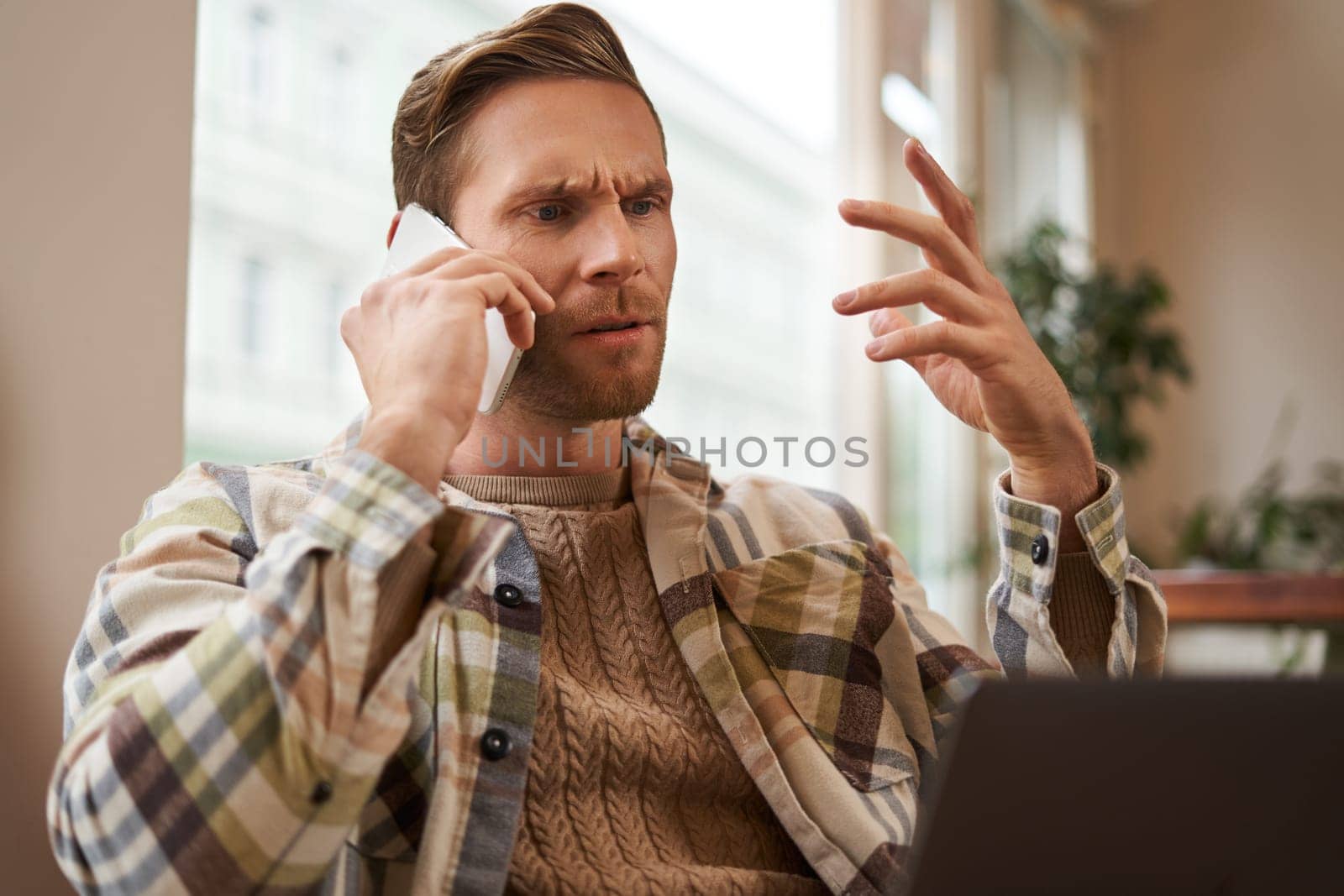 Portrait of confused man arguing with someone over the phone, businessman discuss project, looking at chart on laptop screen, using mobile phone to make a call, staring at monitor with frustration.