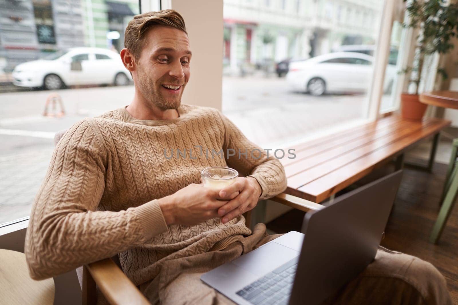 Portrait of handsome modern man, sitting in coffee shop with laptop, looking at screen with happy face, drinking cappuccino, resting in cafe or working by Benzoix