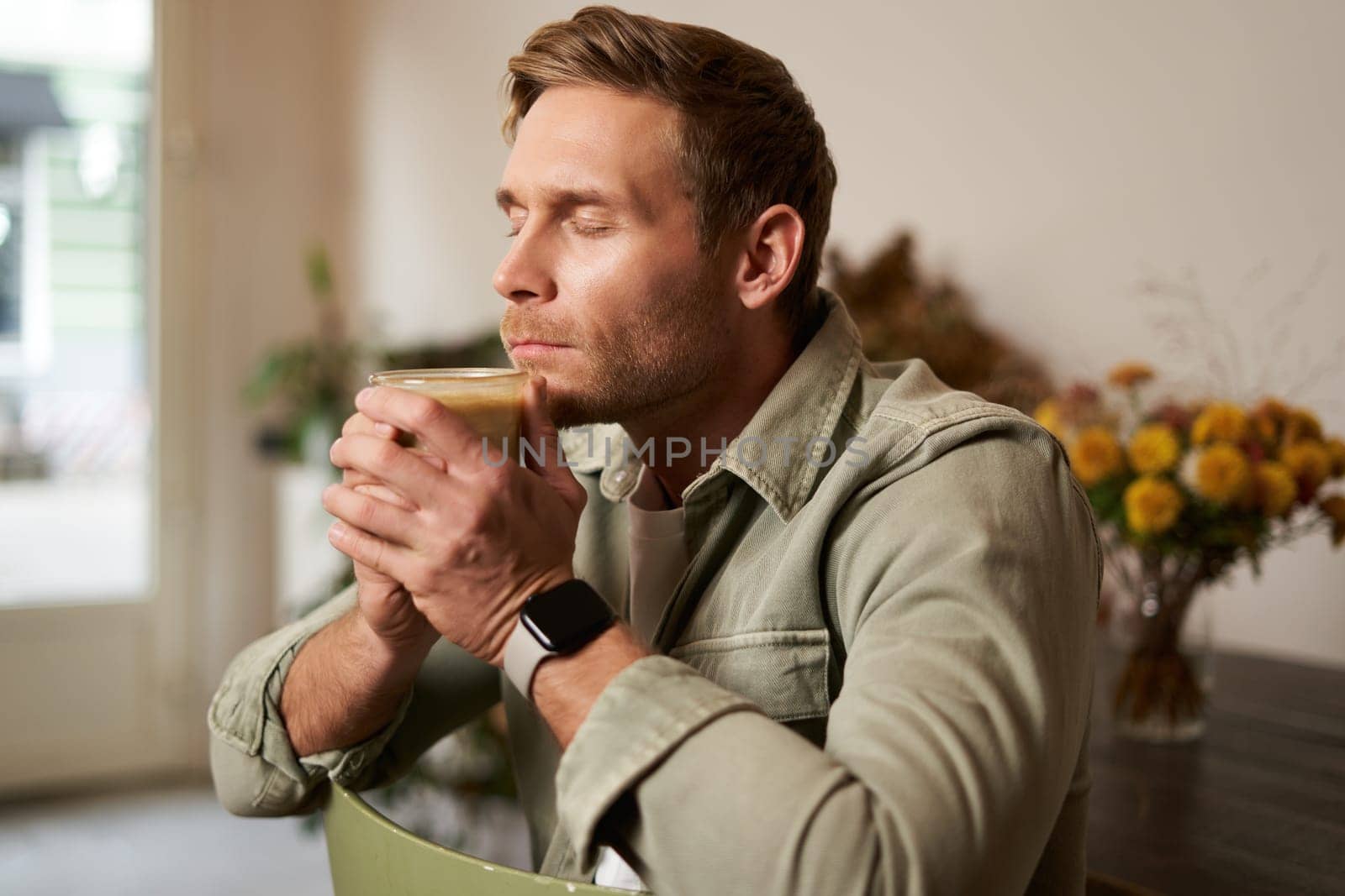 Portrait of young man closes his eyes, smells freshly made coffee in cafe, sits on chair with glass of cappuccino, enjoys the taste.