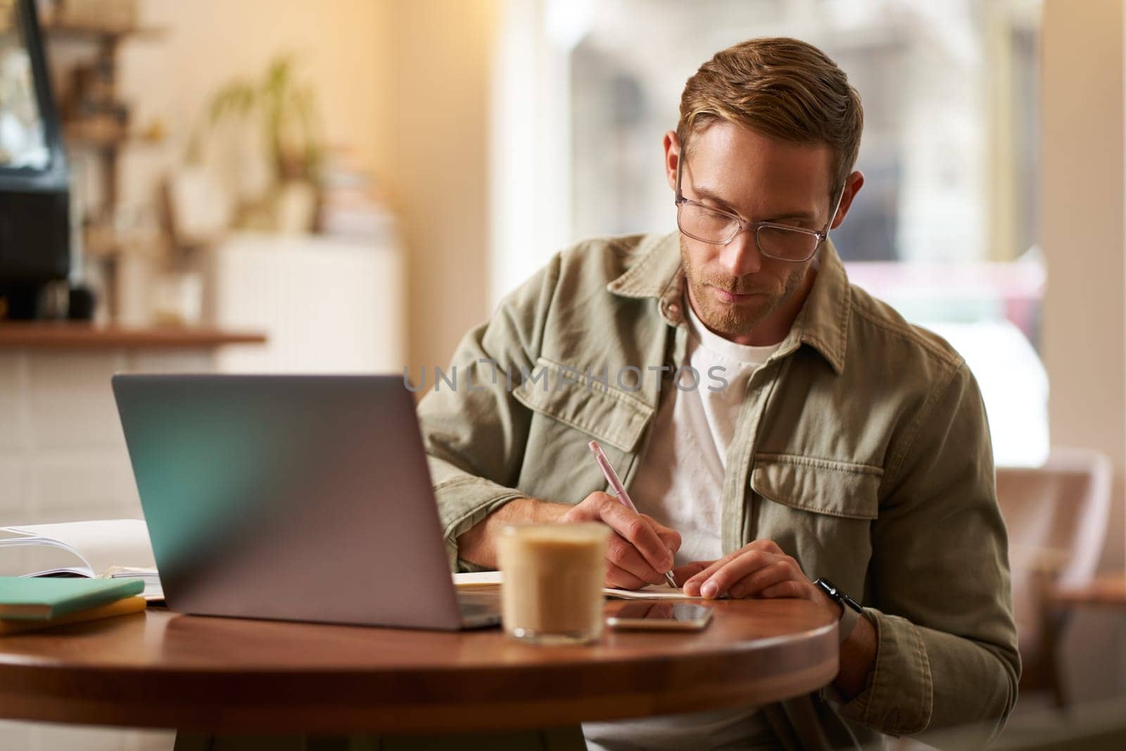 Image of focused young man in glasses, sitting in cafe, making notes, studying, attending online course, learning on remote from quite coffee shop by Benzoix