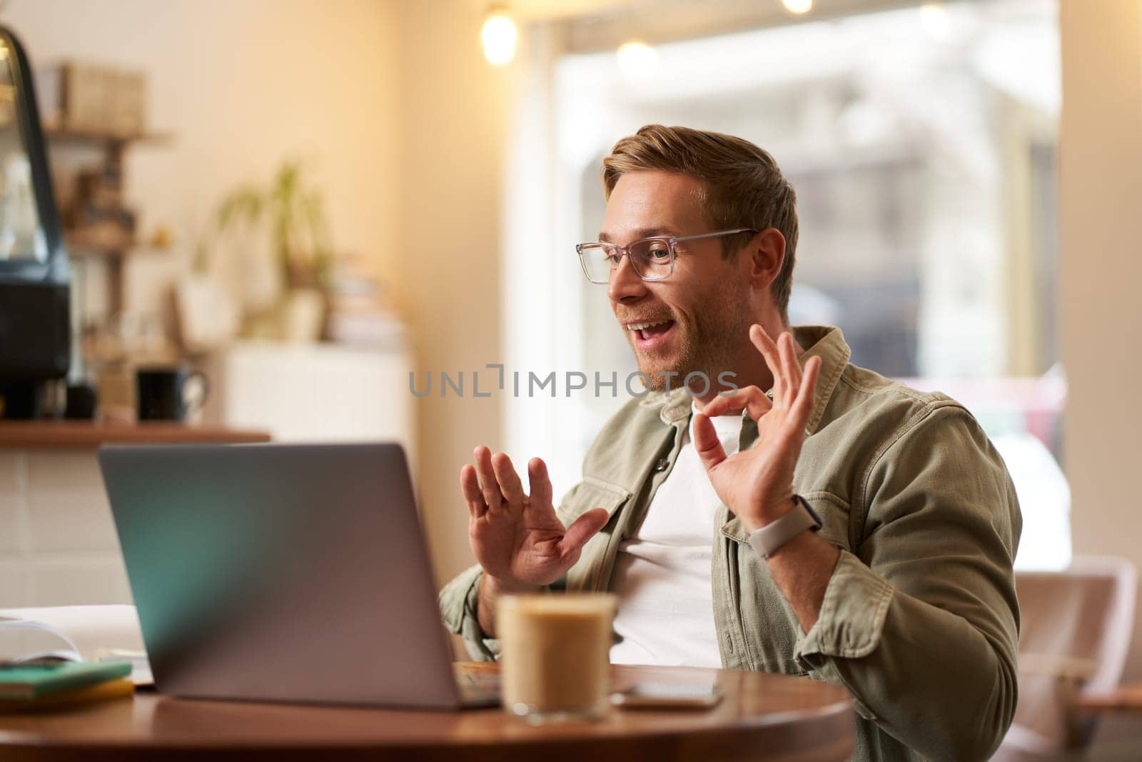 Portrait of young man, teacher, online tutor in glasses, showing okay, ok hand sign, giving lessons remotely from cafe, sitting with laptop, talking to someone on video chat by Benzoix
