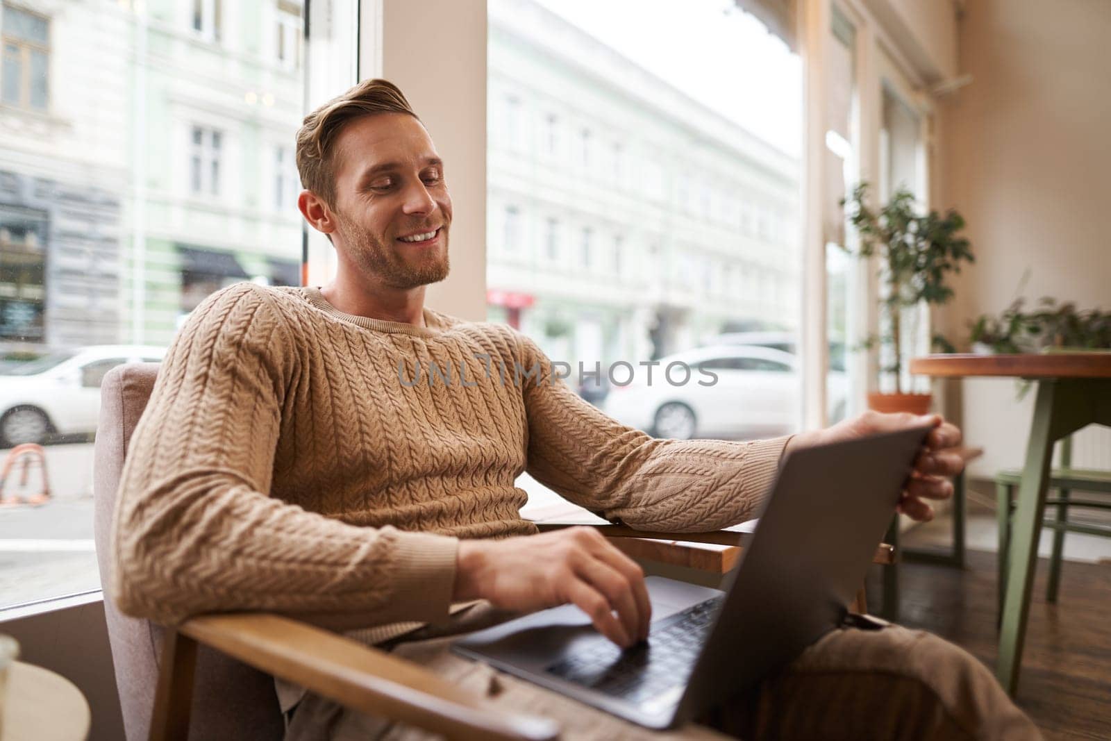 Portrait of young working professional, man sitting in cafe with laptop, drinking coffee and working online, freelancer using coworking space to do his job by Benzoix
