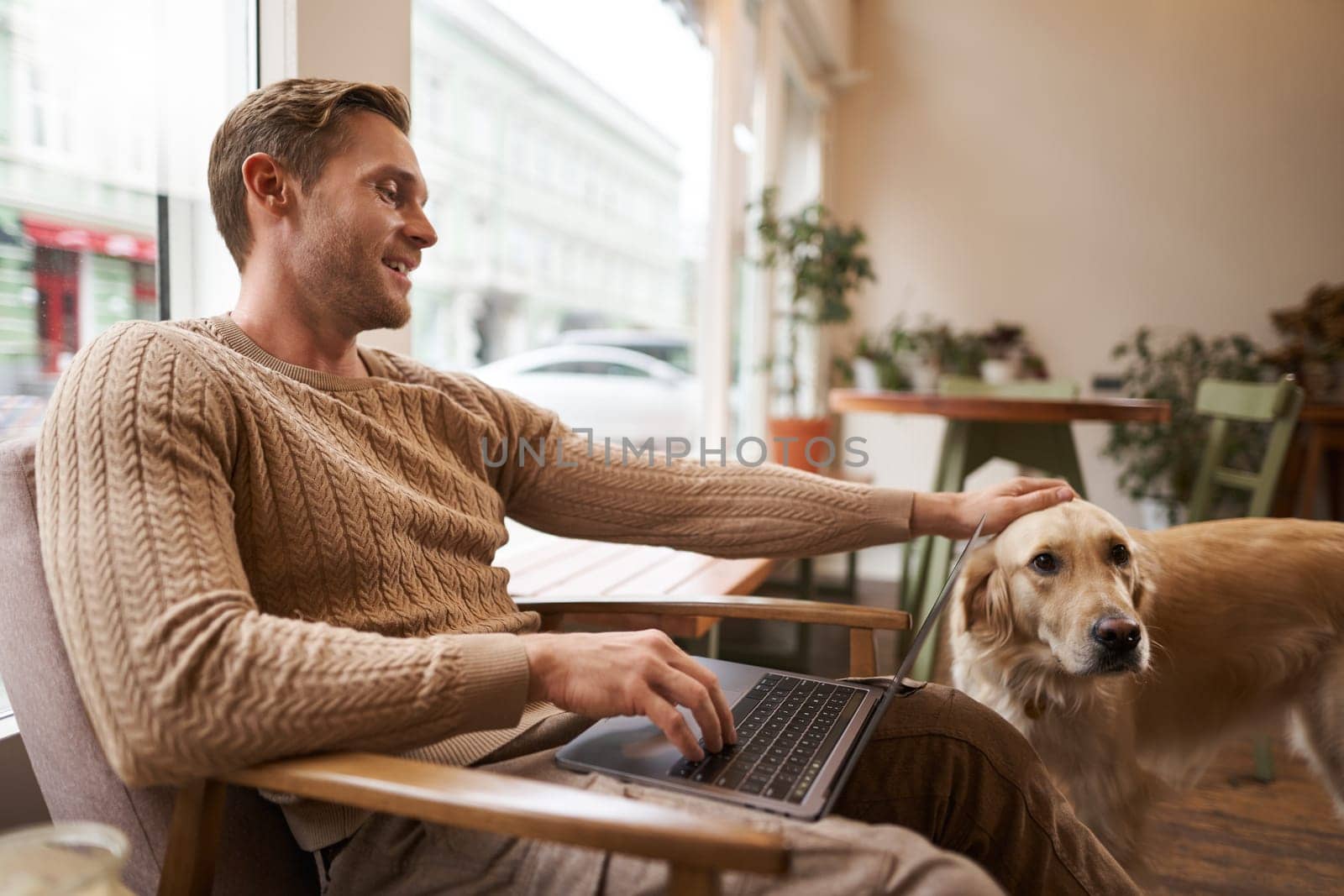 Portrait of young man, digital nomad, sitting in cafe near window, using laptop, working with his pet, petting golden retriever while sitting in chair with computer on laps by Benzoix