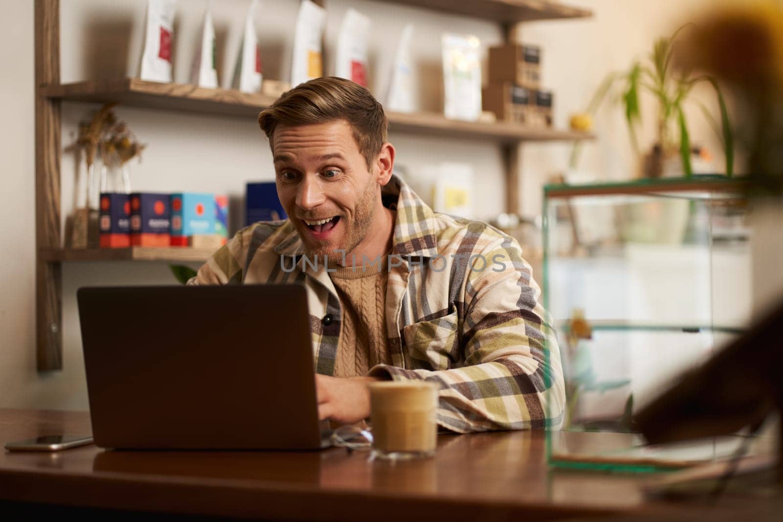 Lifestyle portrait of young businessman, freelancer sitting in cafe, drinking coffee and using laptop, working on online project, digital nomad doing his task, has remote job, looking satisfied.