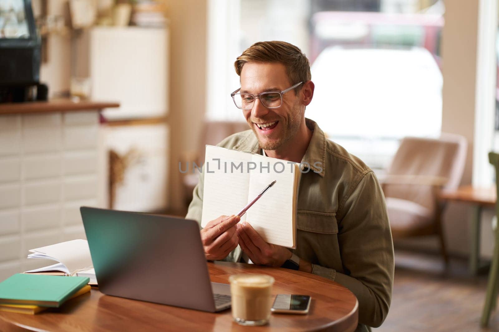 Portrait of young handsome man in glasses, private tutor teaching student online, pointing at his notebook, showing something, video chats via laptop application, working remotely in cafe.