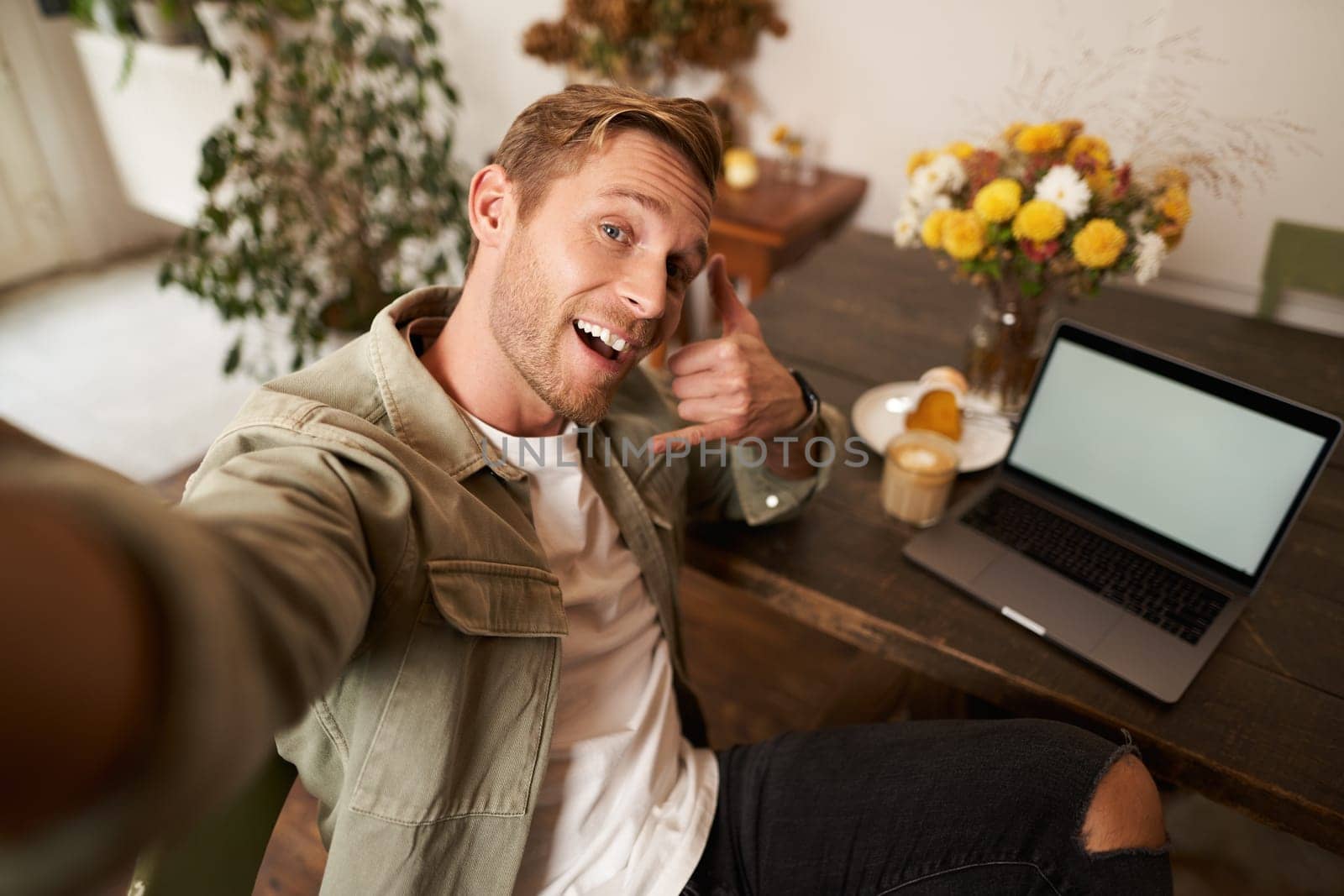 Handsome man takes selfie on his mobile phone with laptop, shows call phone hand sign, smiling at camera, posting photo on social media app, sits in cafe by Benzoix