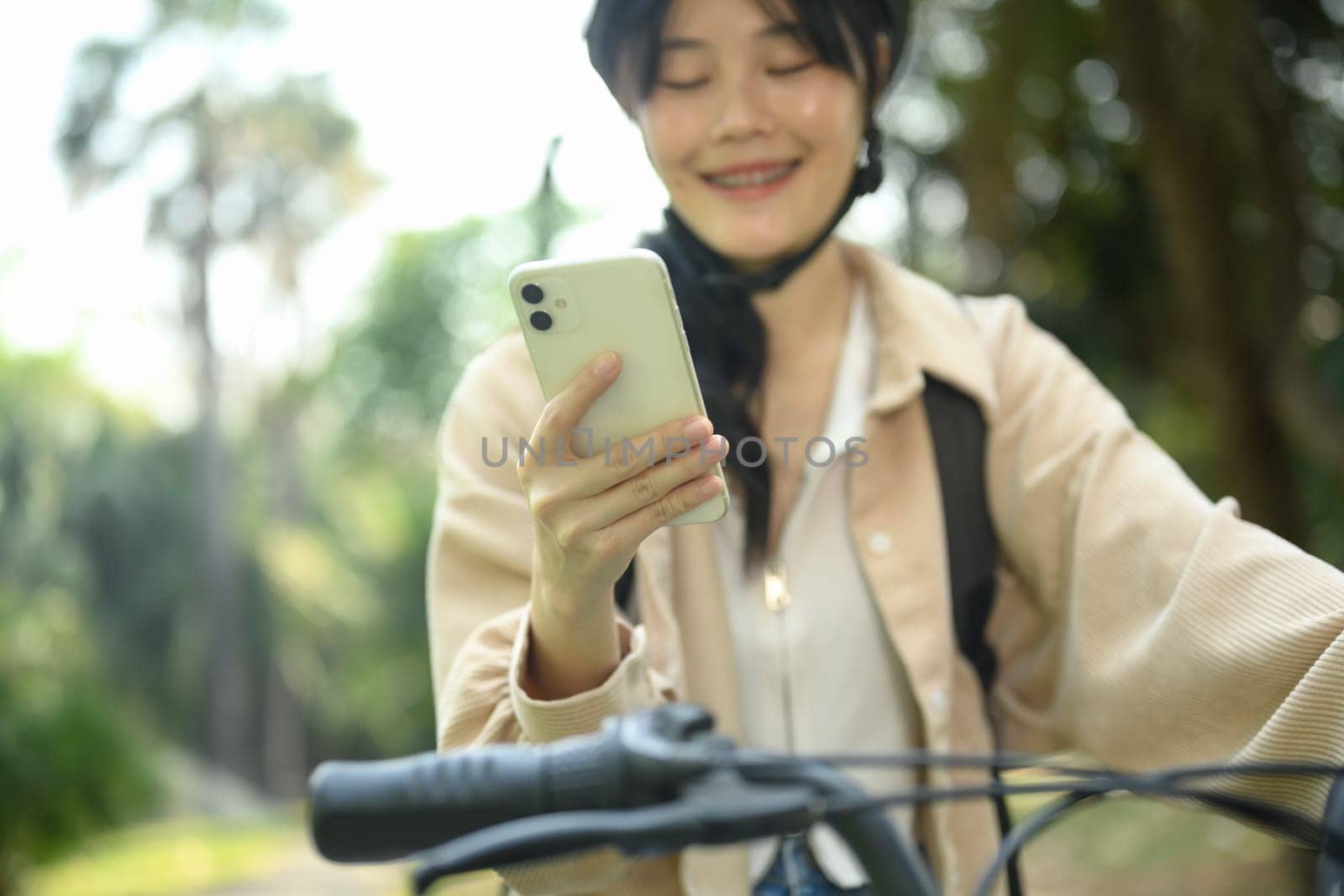 Smiling young Asian woman using mobile phone while standing with her bicycle in public park.