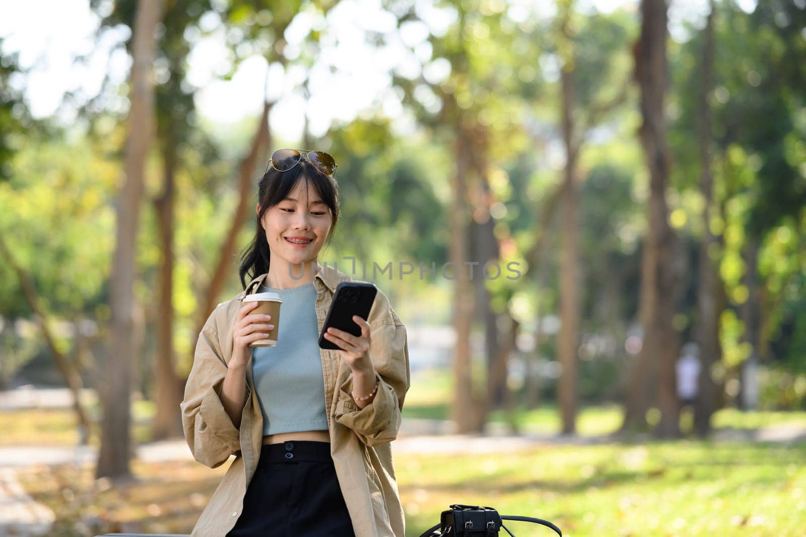 Beautiful young lady sitting park bench drinking coffee and using mobile phone.