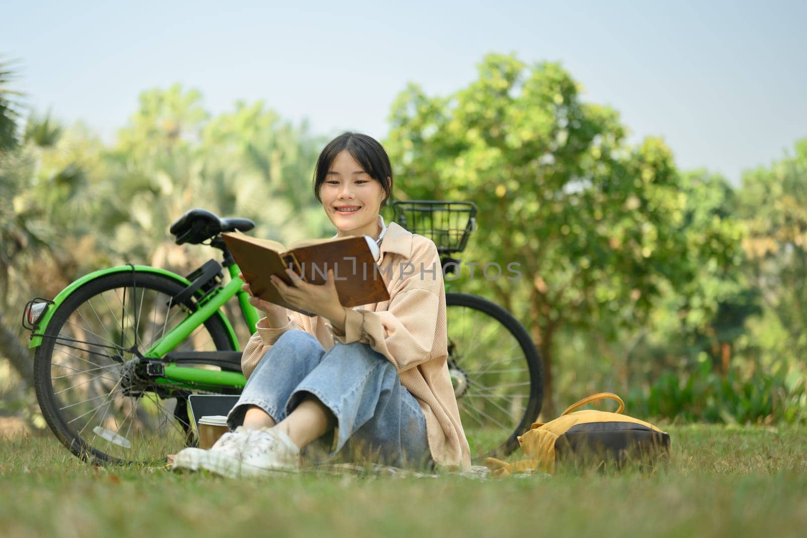 Carefree young woman reading book on green grass at the park with a bicycle.