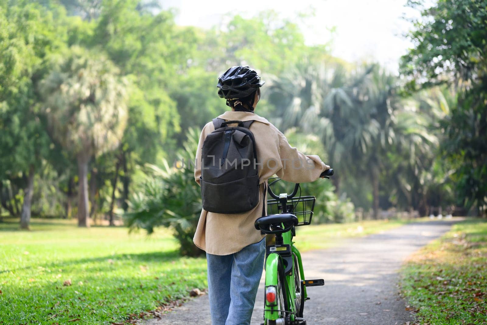 Rear view of young woman walking with bicycle in a sunny park. Lifestyle, transportation, and ECO friendly concept.