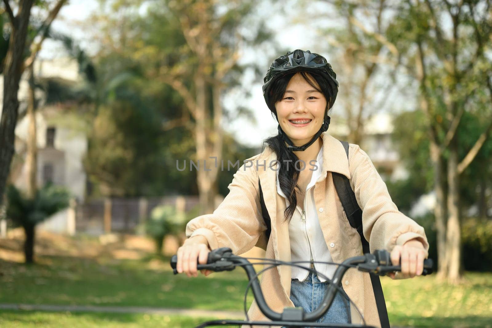 Cheerful young female student in safety helmet riding a bicycle through the city park by prathanchorruangsak