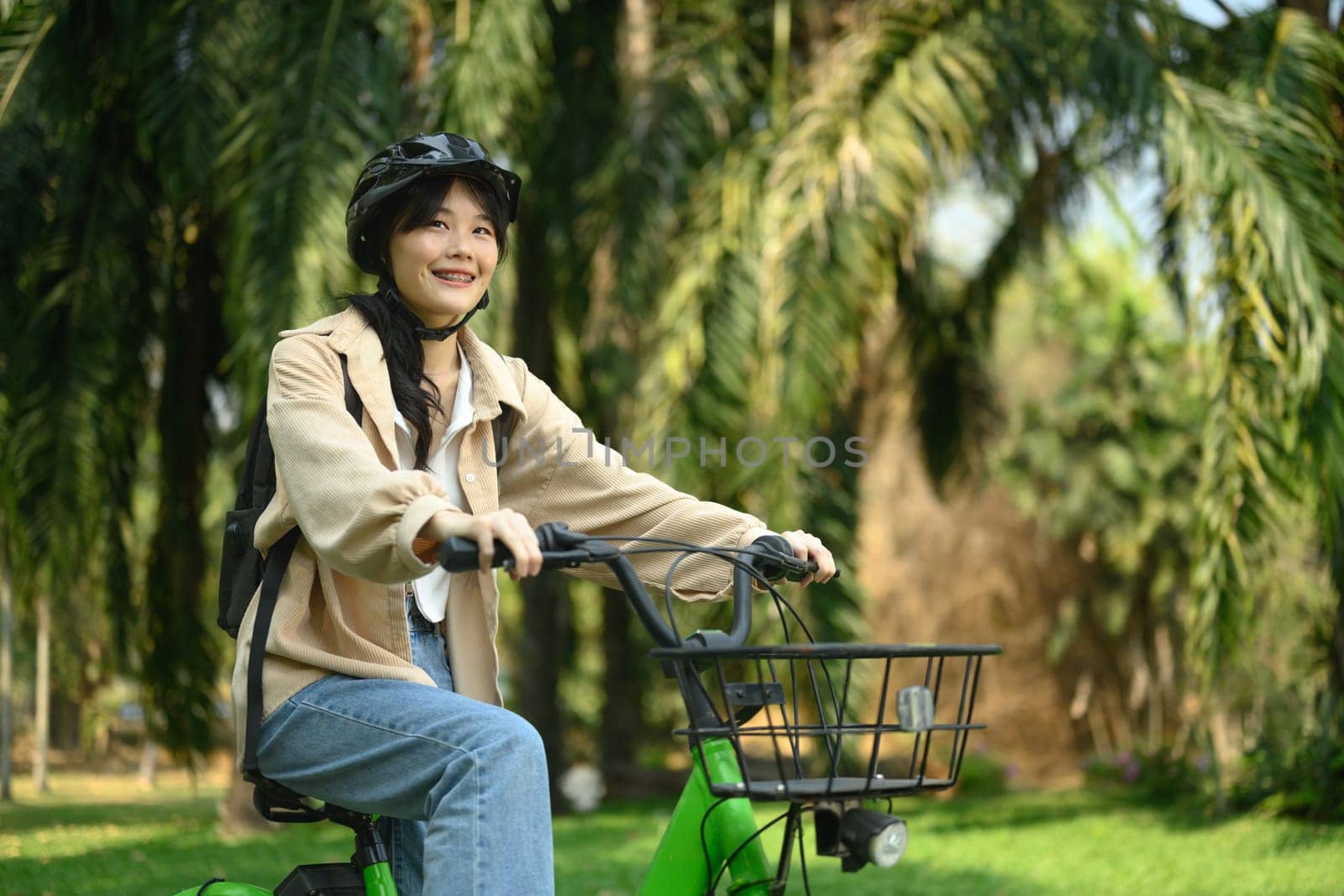 Smiling young woman in safety helmet riding a bicycle through city spring park by prathanchorruangsak