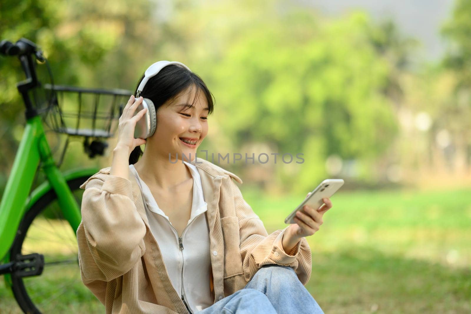 Pretty young woman listening to music with mobile phone and headphones while sitting at the park with a bicycle.