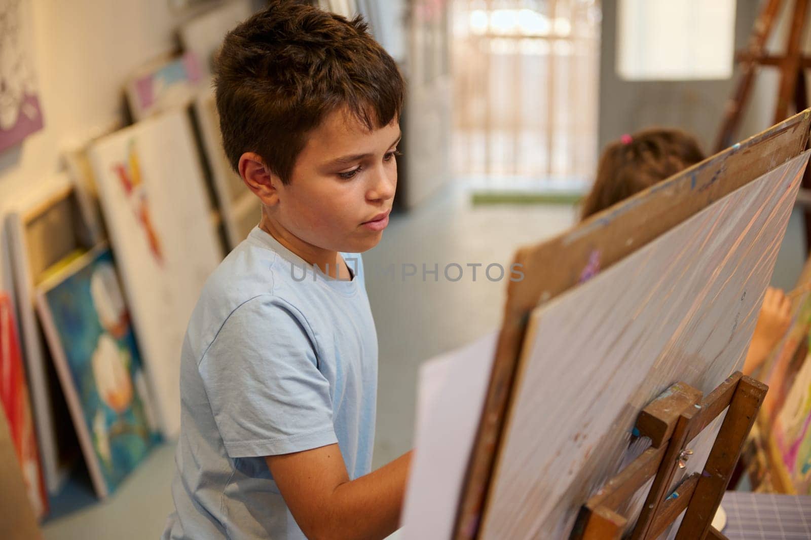 Closeup portrait of an inspired talented boy artist painter painting on a canvas with paintbrush and colorful acrylic paints in a painting class