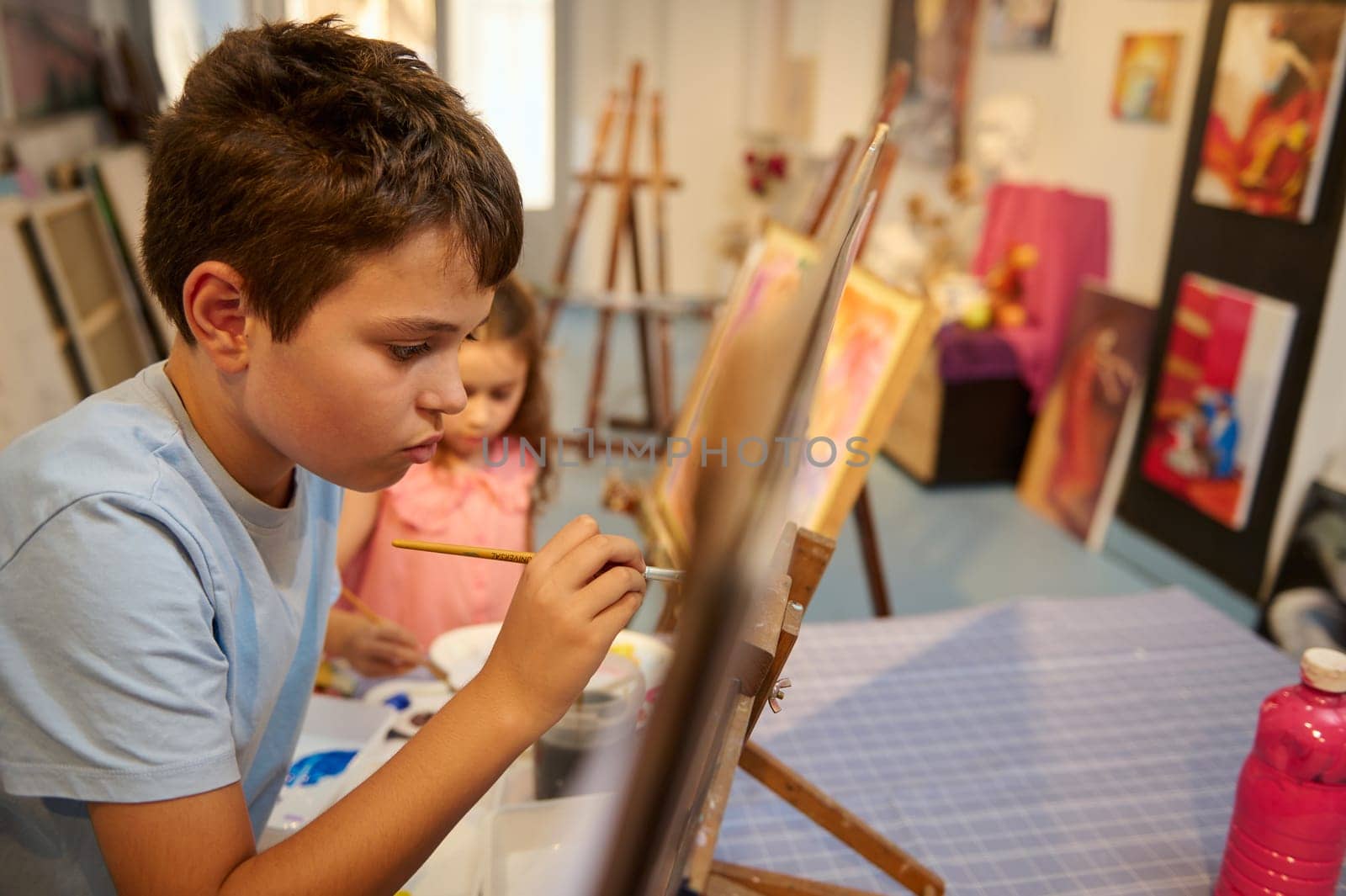 Caucasian handsome teenage school boy focused on painting on canvas in creative art workshop. Acrylic, oil or watercolor painting, drawing, people, kids, education concept