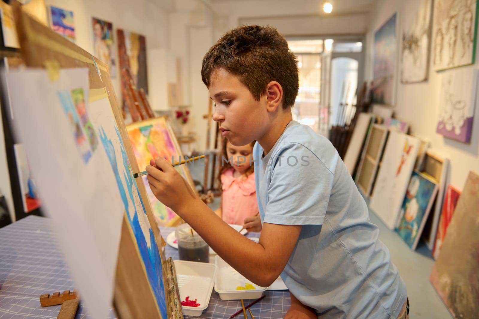 Inspired teenager boy, artist painter painting on canvas in the art school. Talented elementary age kids, boy and girl during fine art, visual art class. Many pictures displayed in the art gallery.