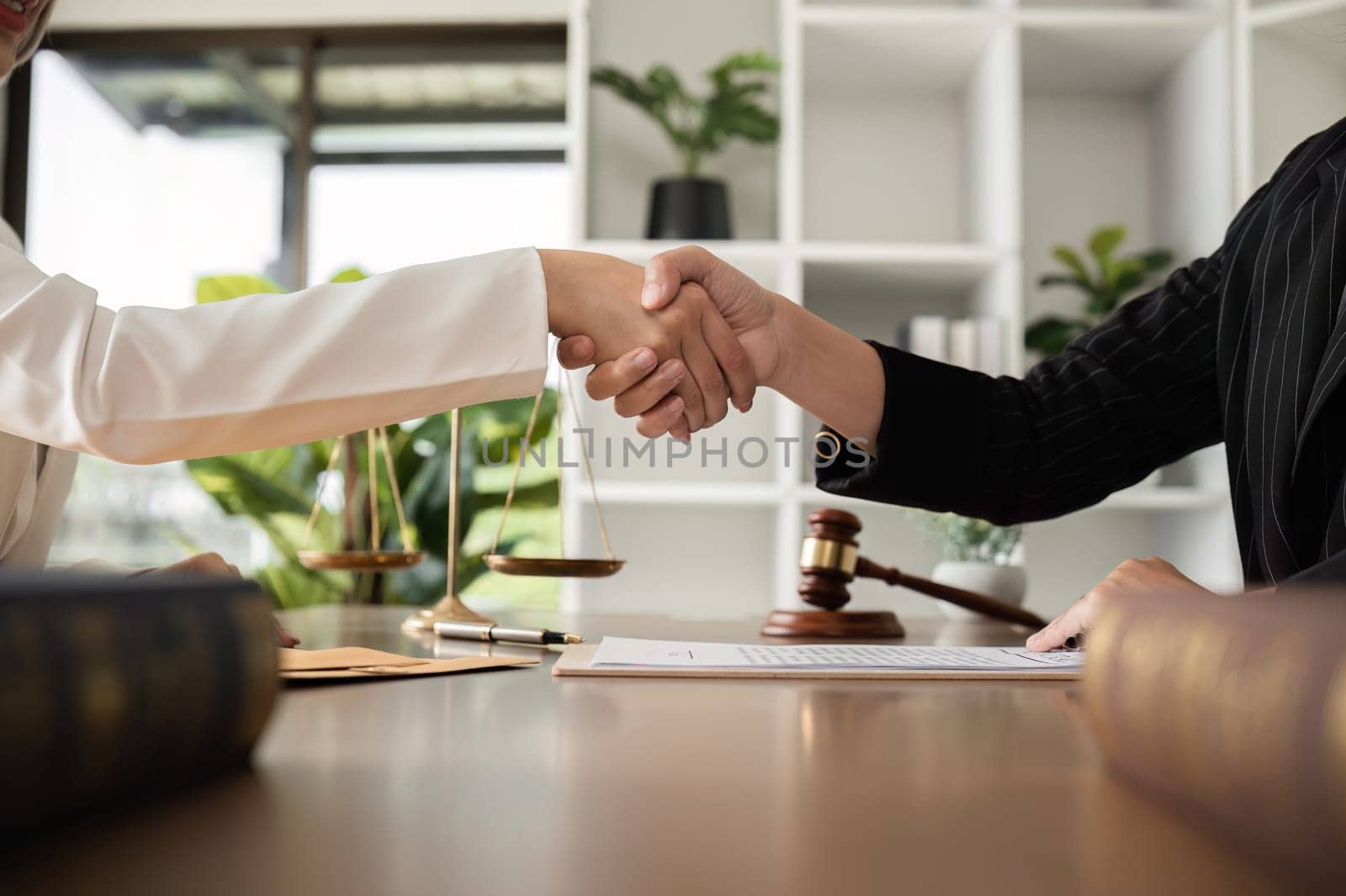 A lawyer and a businesswoman shake hands and reach an agreement on a cooperation contract document. In the lawyer's office and there is a hammer lying on the table. by wichayada