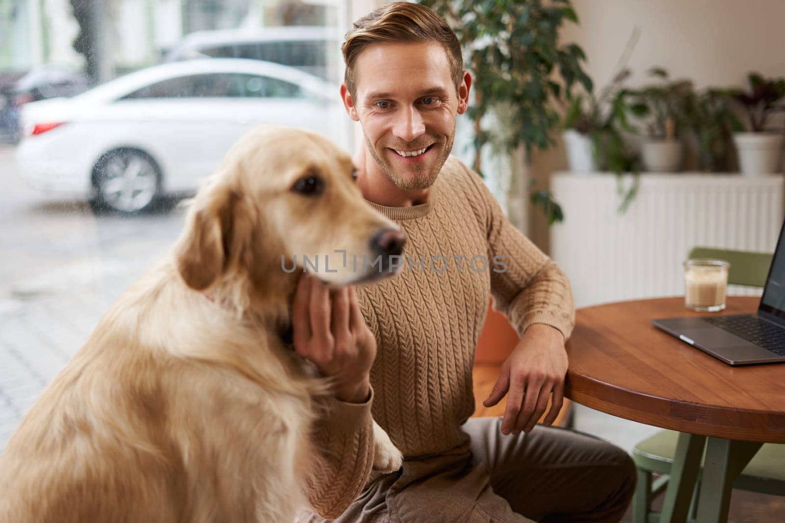 Close up portrait of smiling handsome european man with his dog in a cafe. Guy pets his golden retriever while working outdoors in coffee shop.