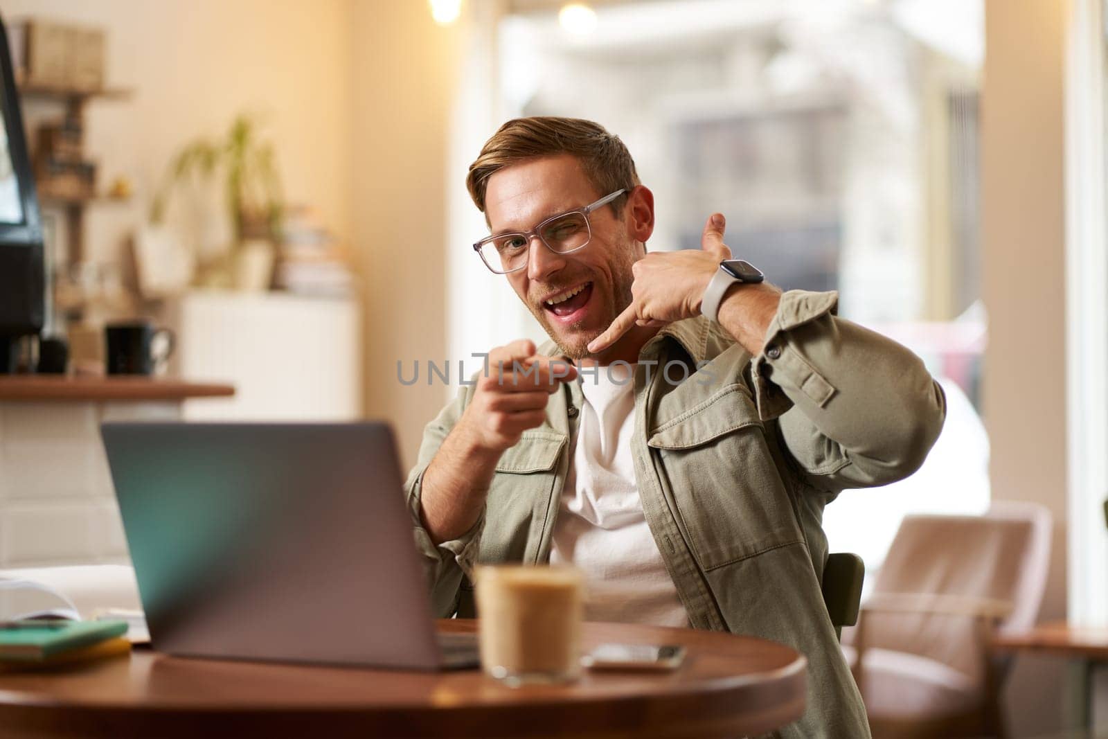 Portrait of cheerful, flirty guy in glasses, sitting with laptop computer in cafe, showing phone call hand gesture and winking at you, giving his number.