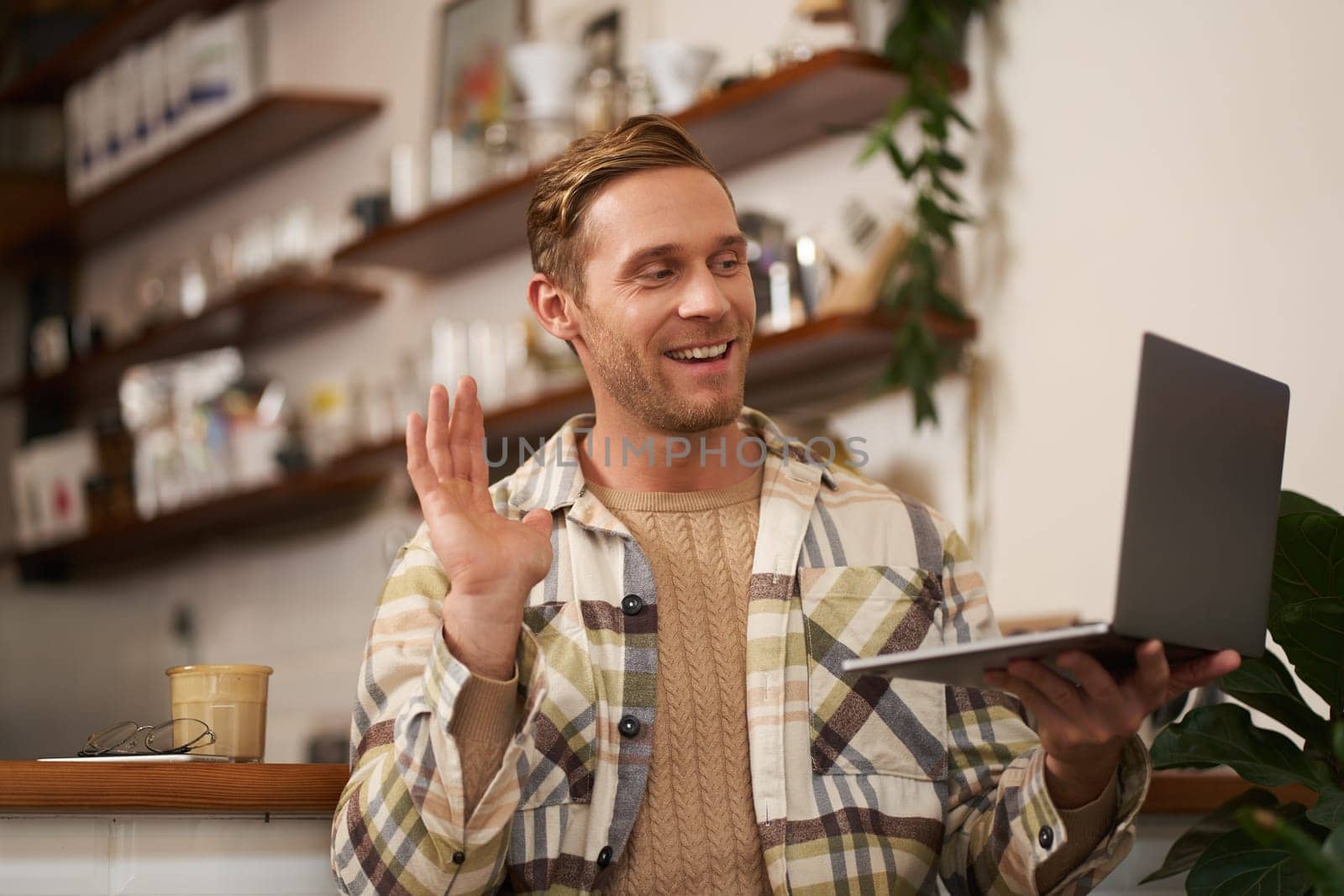 Image of young man in coffee shop, connects to video call, talking online, chats with friends via laptop and waves hand at camera to say hi by Benzoix