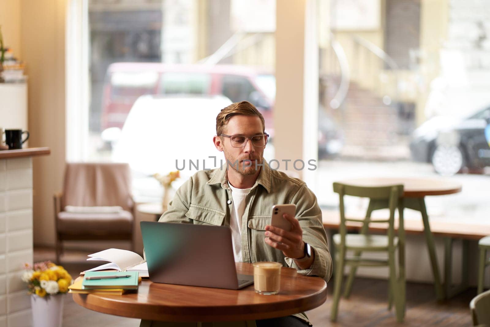 Lifestyle and working people concept. Young man in glasses sitting in empty cafe with laptop and notebook, checking his phone messages, reading on smartphone, working on freelance project.