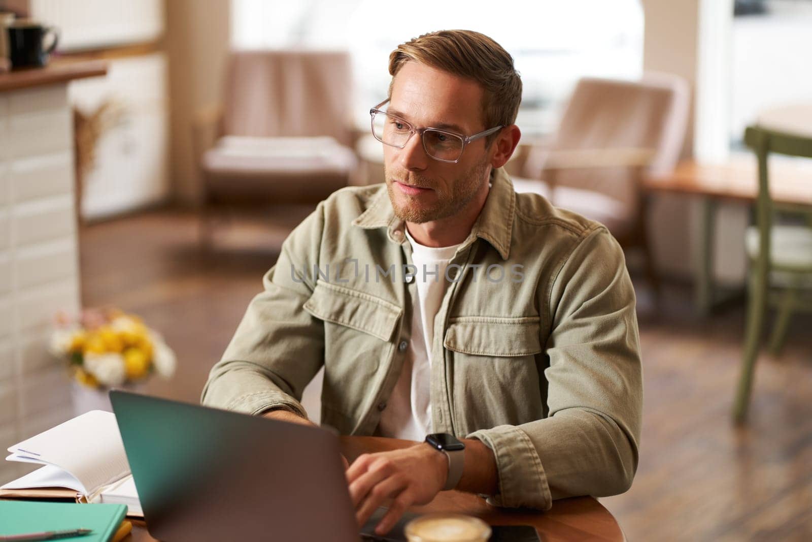 Portrait of young concentrated man in glasses, working from coffee shop, studying, looking thoughtful while completing task on laptop, making notes in notebook.