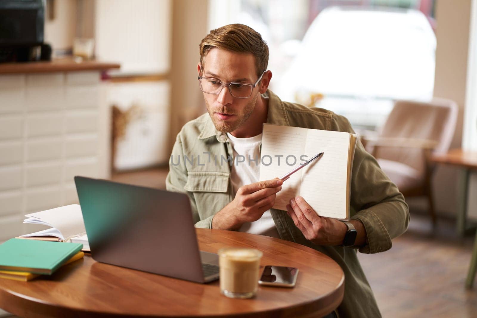 Portrait of handsome man in glasses, working remotely from empty cafe, showing example in his notebook, tutoring, using laptop to meet with student online.