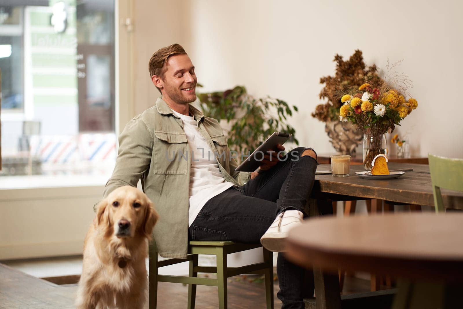 Portrait of handsome young man sits in cafe with his dog, petting golden retriever, reading news on tablet, relaxing with cup of coffee.