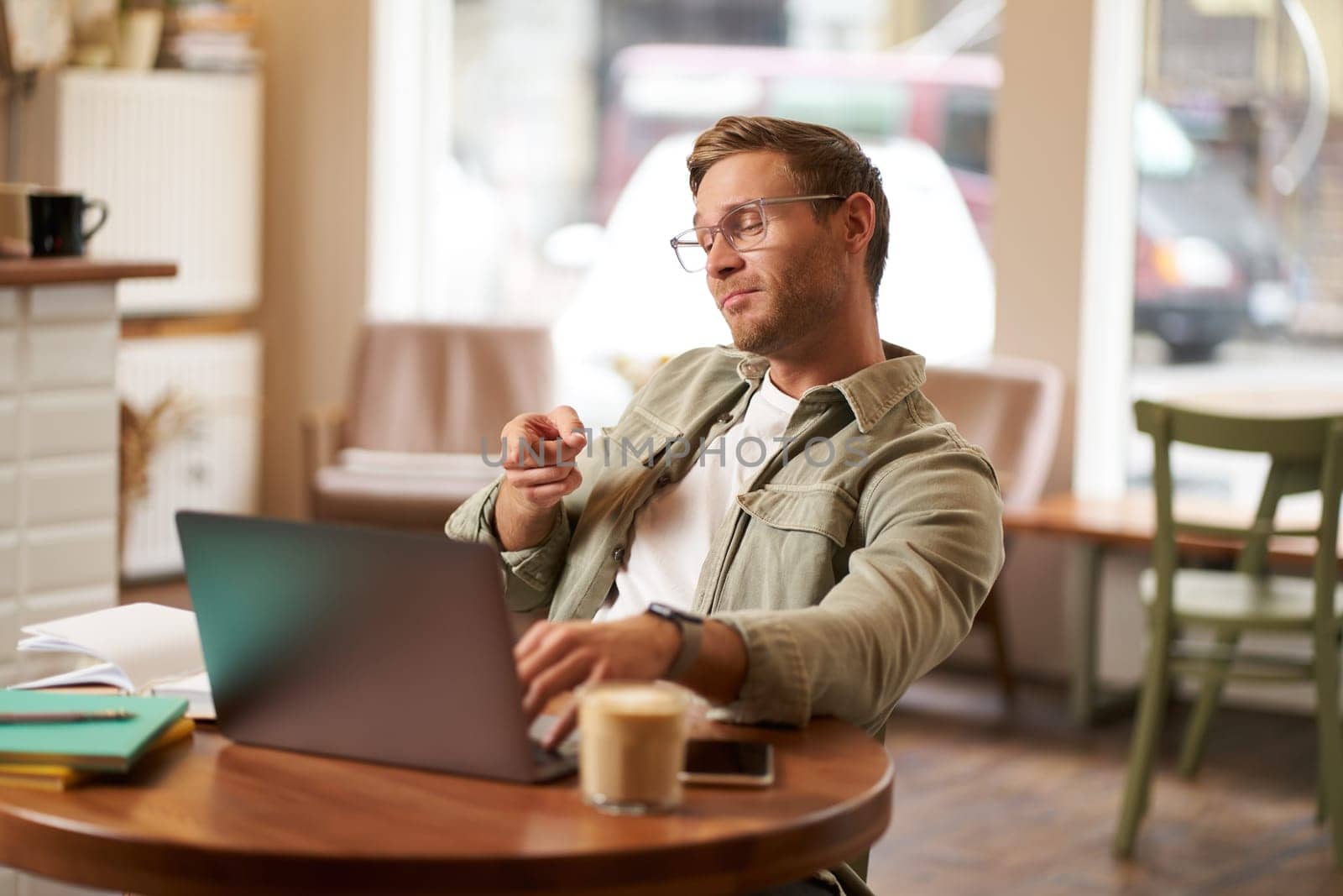Portrait of handsome man in glasses, sits in cafe, points finger at laptop screen, video chats, connects to online meeting, feeling confident, working remotely.
